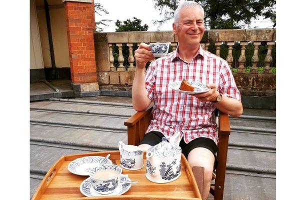 A man smiles whilst holding an Old Chelsea mug up with tea inside. He is sat on a rooftop terrace.