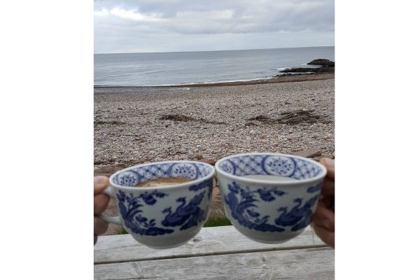 Two people holding Old Chelsea mugs with tea inside. Their view is of the sea with a pebbled beach.