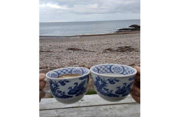 Two people holding Old Chelsea mugs with tea inside. Their view is of the sea with a pebbled beach.