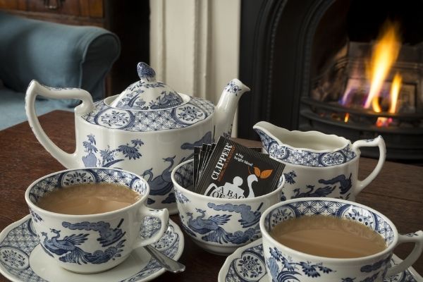 An Old Chelsea patterned teapot and two mugs sit on a tray in front of a fire. Clipper teabags are on the tray.