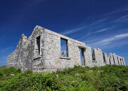 Grey brick ruin of a single-storey building with no roof on the edge of a cliff with views of the sky beyond